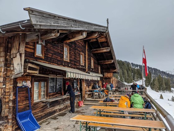 Trattenbachalm im Winter Rustikale Holzhütte mit Außenterrasse, Gästen an Biertischen und einer österreichischen Flagge im Schnee.