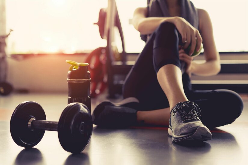 Fitnesspause im Trainingsraum Person sitzt am Boden eines Fitnessraums neben einer Hantel und einer Trinkflasche.