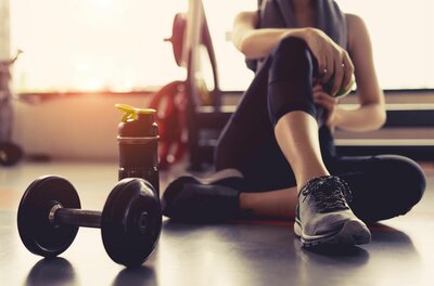 Person sitzt am Boden eines Fitnessraums neben einer Hantel und einer Trinkflasche.