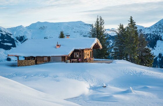 Schneebedeckte Holzhütte in einer alpinen Winterlandschaft mit Bergen im Hintergrund.