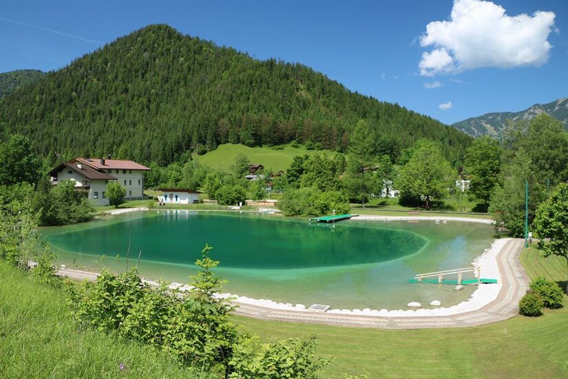 Ein Genuss für Wasserratten! Ein ruhiger See umgeben von grünen Hügeln und dichten Wäldern. Im Hintergrund sind ein paar Häuser und ein klarer blauer Himmel sichtbar.