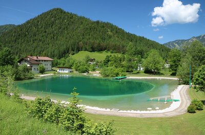 Ein ruhiger See umgeben von grünen Hügeln und dichten Wäldern. Im Hintergrund sind ein paar Häuser und ein klarer blauer Himmel sichtbar.