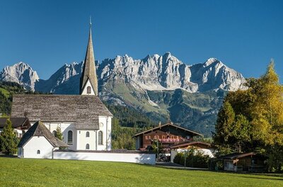 Weiße Kirche mit spitzem Turm vor einem massiven Gebirgszug unter blauem Himmel.