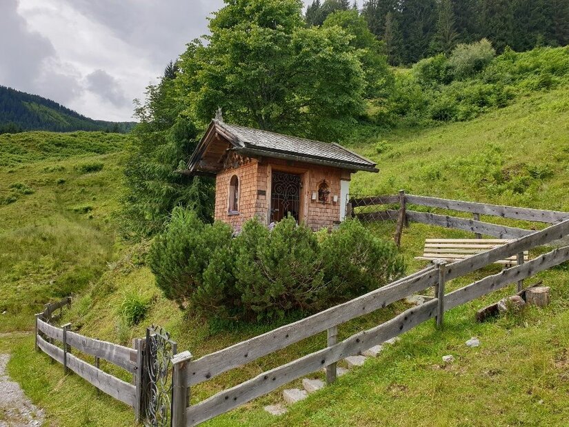Kleine Holzkapelle mit Schindelfassade auf einer grünen Almwiese hinter einem Holzzaun.