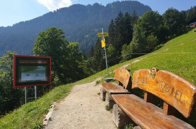 Rastplatz am Alten Seidlalmweg Wanderweg mit Holzbank, Infotafel und Wegweisern vor bewaldetem Berghang.