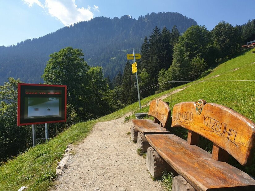 Rastplatz am Alten Seidlalmweg Wanderweg mit Holzbank, Infotafel und Wegweisern vor bewaldetem Berghang.