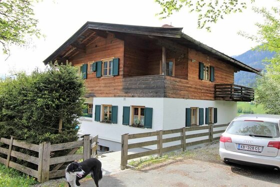 Lodge Lebenberg Zweistöckiges Holzhaus mit weißem Erdgeschoss, grünen Fensterläden und einem davor stehenden Hund.