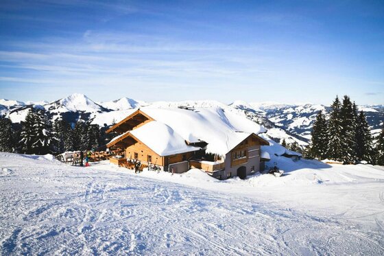Schneebedeckte Almhütte aus Holz in einer alpinen Winterlandschaft mit Bergen im Hintergrund.