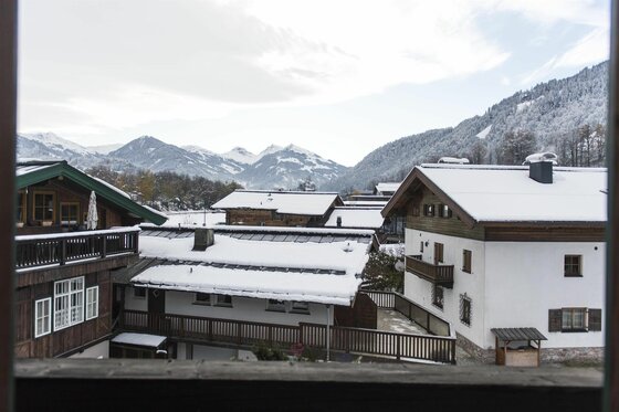 Aussicht vom Apartment Wilder Kaiser Verschneite Hausdächer in einem Alpendorf vor bewaldeten Bergen und schneebedeckten Gipfeln.