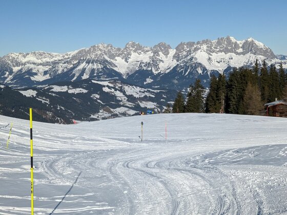 Präparierte Skipiste im Vordergrund mit Blick auf ein verschneites Gebirgsmassiv unter blauem Himmel.