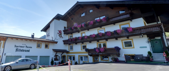 Mehrstöckiges Gebäude der Apartment-Pension Hillebrand mit Holzbalkonen und Blumenschmuck unter blauem Himmel.