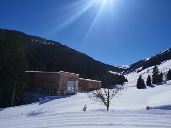 Modernes Holzgebäude in einer verschneiten Gebirgslandschaft unter blauem Himmel mit Sonneneinstrahlung.