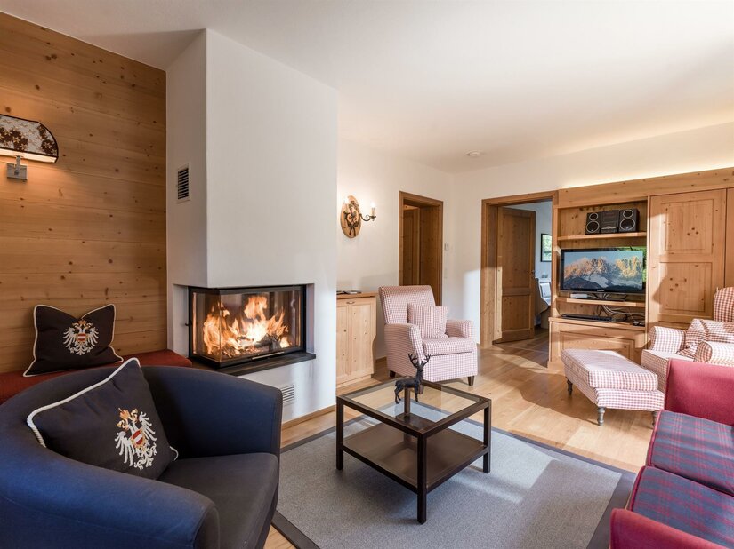 Wohnzimmer mit brennendem Kamin, Sesseln, Couchtisch und Fernseher in alpiner Holzoptik.