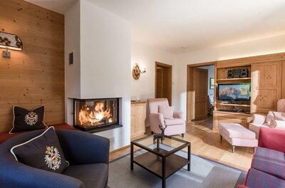 Wohnzimmer mit brennendem Kamin, Sesseln, Couchtisch und Fernseher in alpiner Holzoptik.