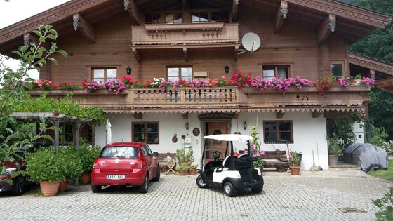 Alpines Holzhaus mit blumengeschmücktem Balkon, davor ein rotes Auto und ein weißes Golfcart auf gepflastertem Vorplatz.