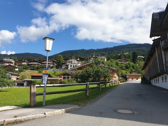 Siedlung am Rauhen Kopf Asphaltierte Straße in einer Siedlung mit Holzhäusern vor bewaldeten Bergen unter blauem Himmel.