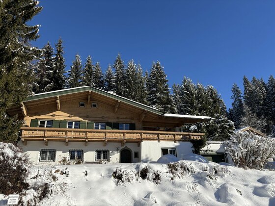 Alpines Holzhaus mit Balkon und grünen Fensterläden in einer verschneiten Winterlandschaft vor Nadelwald.