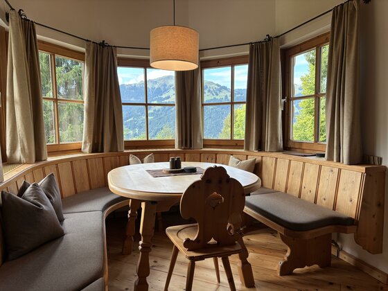 IMG_1898 Ein gemütlicher Essbereich mit einem runden Tisch und Holzbänken. Große Fenster bieten einen Blick auf die Berge und die Natur.