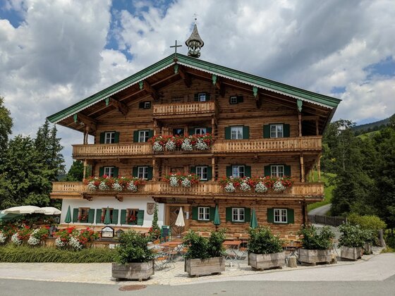 Alpengasthof mit Außenterrasse Mehrstöckiges traditionelles Holzgebäude mit Blumenbalkonen und Außengastronomie unter bewölktem Himmel.
