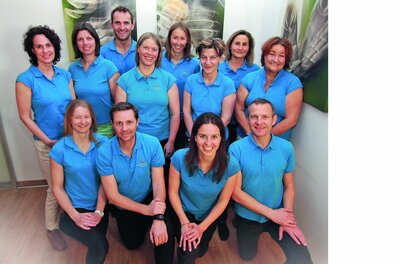Team Balance Physiotherapie Ein elfköpfiges Team in blauen Poloshirts posiert in einem hellen Innenraum für ein Gruppenfoto.