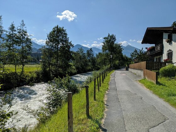 Uferweg an der Kitz-Connection Asphaltierter Weg neben einem fließenden Bach mit Holzzaun, Bäumen und Bergen im Hintergrund.