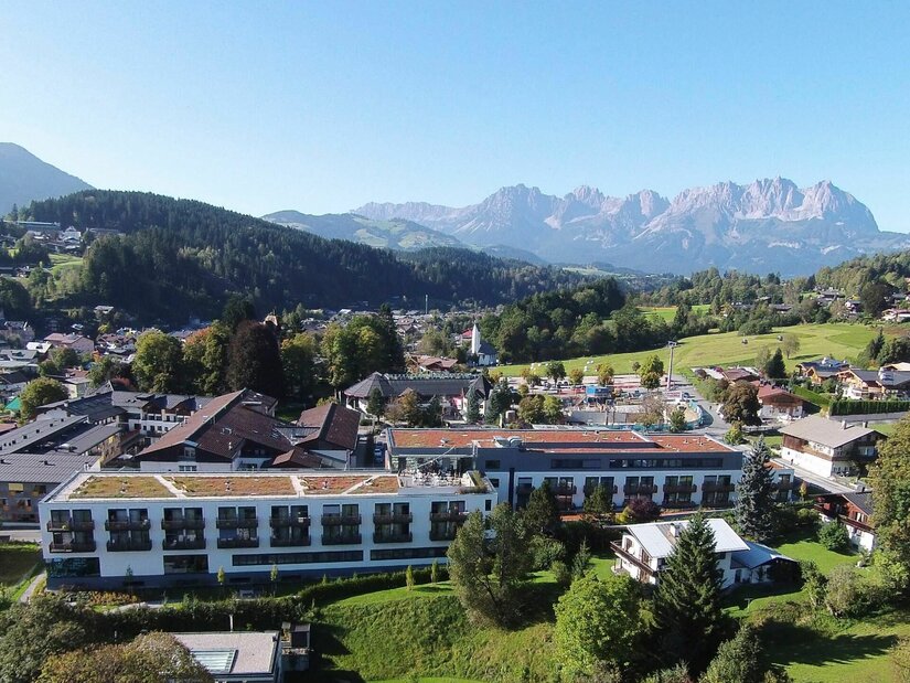 Rehazentrum Kitzbühel im Sommer Luftaufnahme eines modernen Klinikgebäudes in Kitzbühel vor bewaldeten Hügeln und dem Wilden Kaiser.