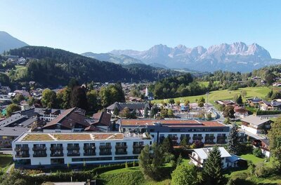 Luftaufnahme eines modernen Klinikgebäudes in Kitzbühel vor bewaldeten Hügeln und dem Wilden Kaiser.
