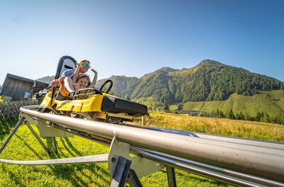 Eine aufregende Sommerrodelbahn in den Bergen. Zwei Personen genießen die Fahrt bei schönem Wetter und klarem Himmel.