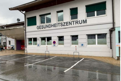 Ein Gesundheitszentrum mit einer modernen Fassade und großen Fenstern. Der Parkplatz ist teilweise leer und die Straße ist nass von Regen.