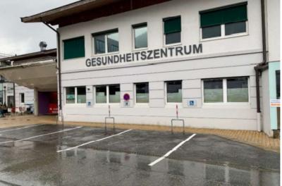 Ein Gesundheitszentrum mit einer modernen Fassade und großen Fenstern. Der Parkplatz ist teilweise leer und die Straße ist nass von Regen.