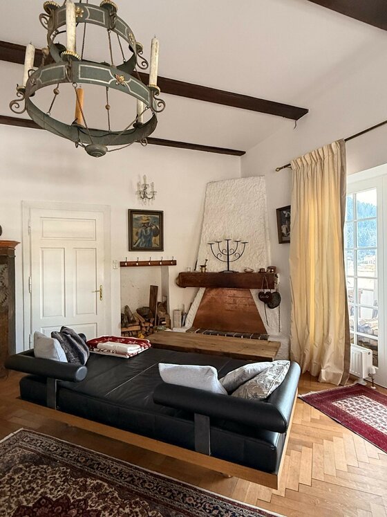 Ein gemütliches Wohnzimmer mit einem modernen Sofa und einem rustikalen Kamin. Der Raum hat helle Wände und große Fenster mit Aussicht.