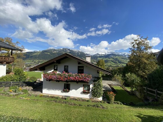 Alpines Wohnhaus mit blumengeschmücktem Balkon vor schneebedeckten Bergen unter blauem Himmel.