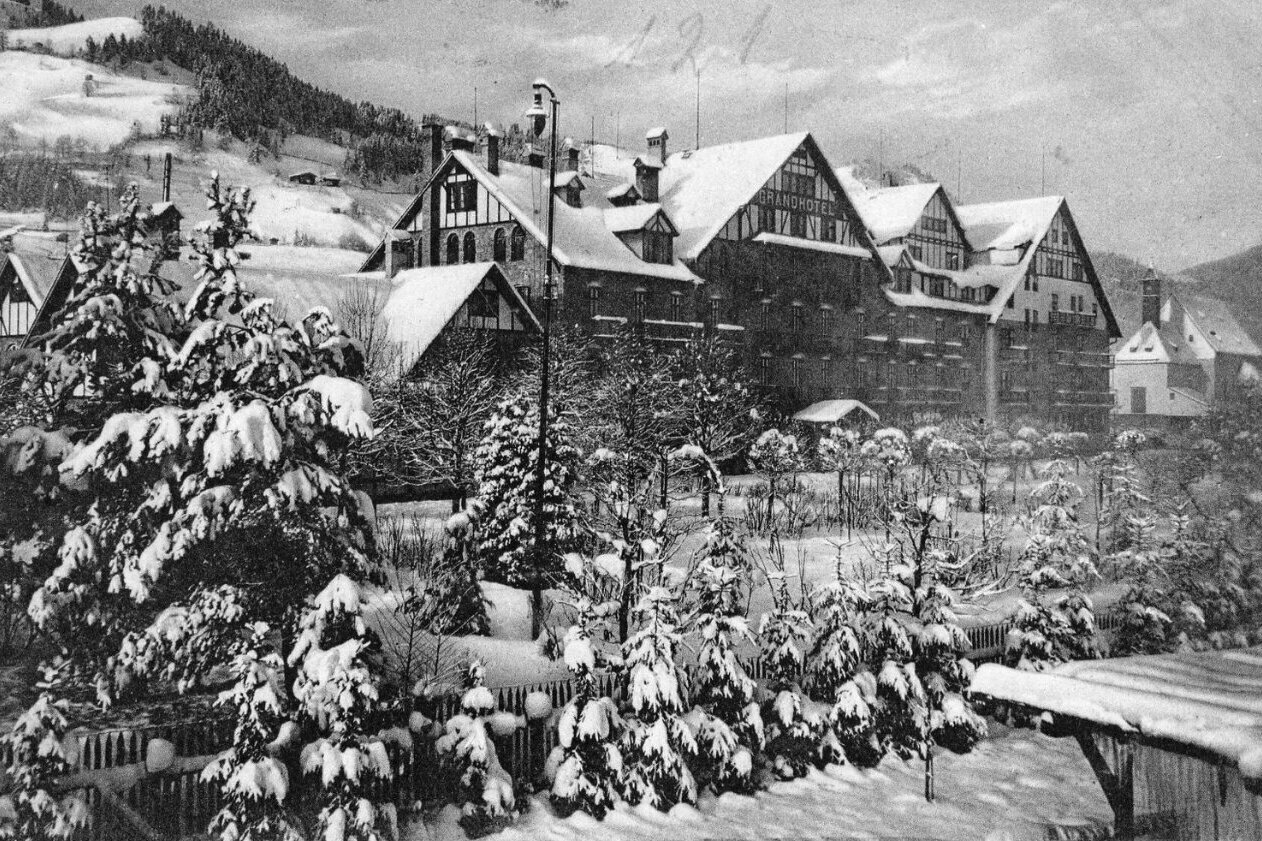 Black-and-white photograph of the Grand Hotel Kitzbühel in winter, with snow-covered trees and bushes