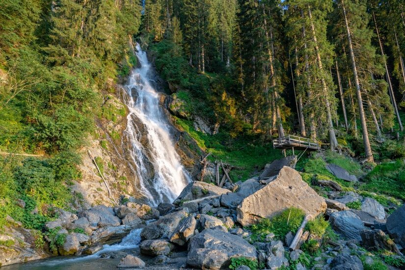 Ein Wasserfall stürzt über Felsen in einem Nadelwald herab, im Vordergrund liegen große graue Steine.