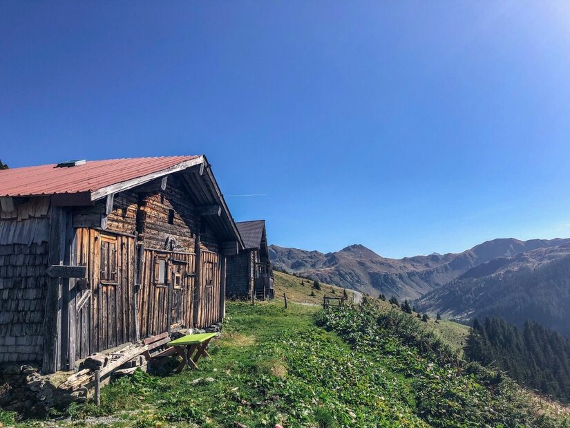 Rustikale Holzhütten auf einer grünen Almwiese vor einem Gebirgspanorama unter blauem Himmel.