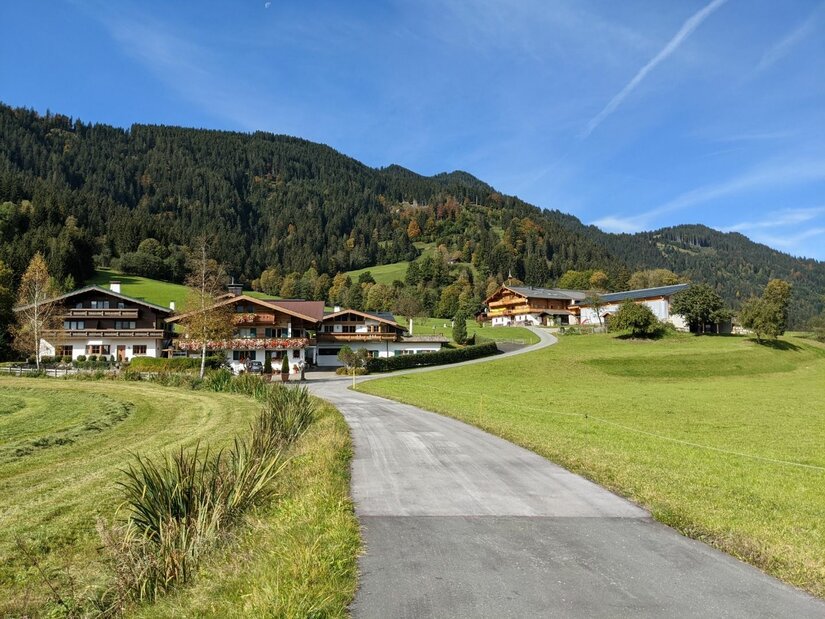 Alpenhäuser bei Reith Asphaltierter Weg führt zu einer Gruppe von Alpenhäusern vor bewaldeten Bergen unter blauem Himmel.
