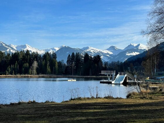 Blick über einen See auf eine Badeanlage mit Rutsche und Sprungturm vor verschneiten Berggipfeln.