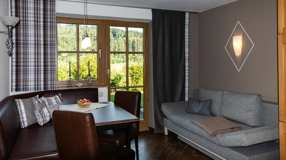 Wohnzimmer Ferienwohnung Hauser Wohnbereich mit Eckbank, Holztisch und grauem Sofa vor einem Fenster mit Blick ins Grüne.