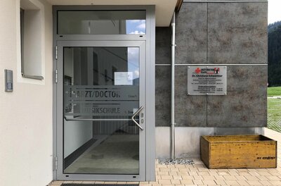 Eingangsbereich einer Arztpraxis mit Glastür und Hinweisschild an einer grauen Fassade.