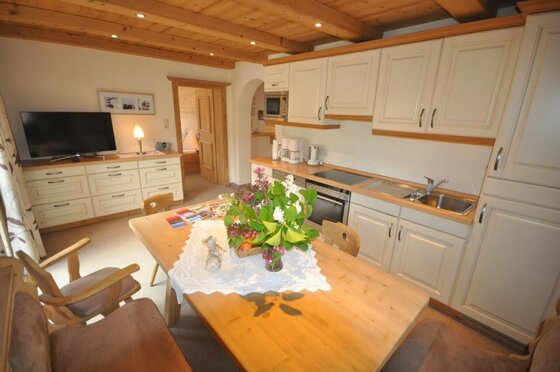 Helle Wohnküche mit Holztisch, Küchenzeile, Fernseher und Holzdecke.