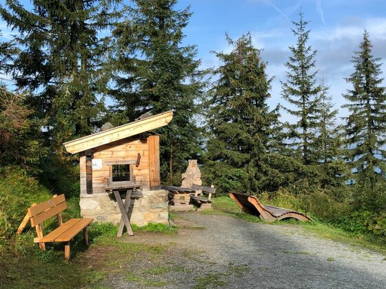 Kleine Holzhütte mit Pultdach, Sitzbänken und einer Holzliege an einem Schotterweg vor Nadelbäumen.