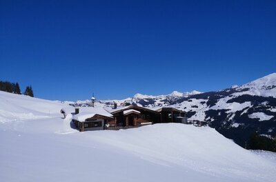Holzhütte in einer verschneiten Gebirgslandschaft unter blauem Himmel.