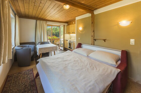 Innenansicht einer Ferienwohnung mit Doppelbett, Sitzecke, kleiner Küchenzeile und Balkonzugang.