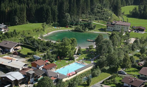 Überflug Badeanlage Ein malerischer Ort mit einem klaren See und einem Schwimmbecken. Umgeben von grünen Wäldern und gemütlichen Häusern.