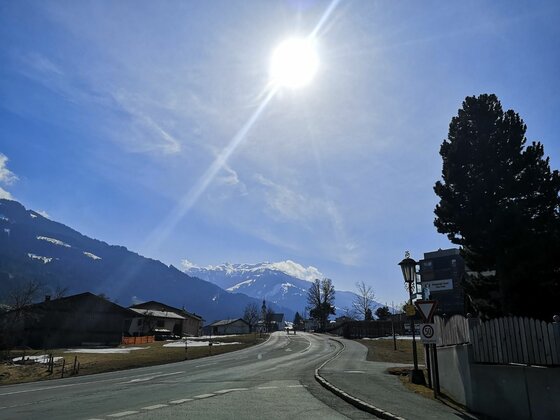 Asphaltierte Straße in einer Siedlung mit Blick auf schneebedeckte Berge unter strahlender Sonne.