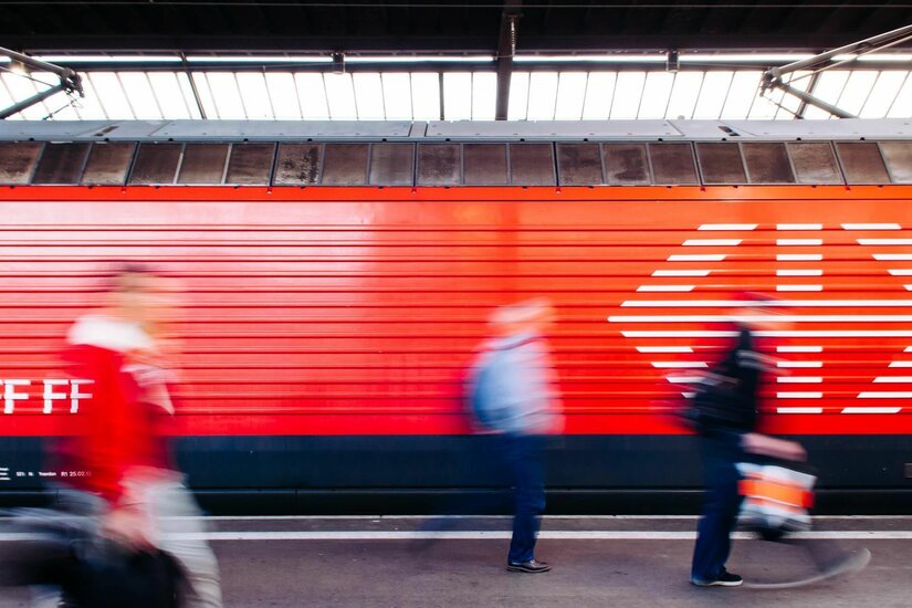 Seitenansicht eines roten Zuges am Bahnsteig mit vorbeilaufenden, unscharfen Personen.