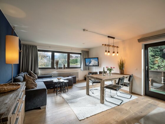 Modernes Wohnzimmer mit grauem Ecksofa, Holzesstisch und Blick durch ein Fenster auf die Berge.