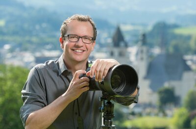 Markus Mitterer Fotografie Ein lächelnder Fotograf mit einer Kamera steht im Freien. Im Hintergrund sind unscharfe Gebäude und eine grüne Landschaft zu sehen.