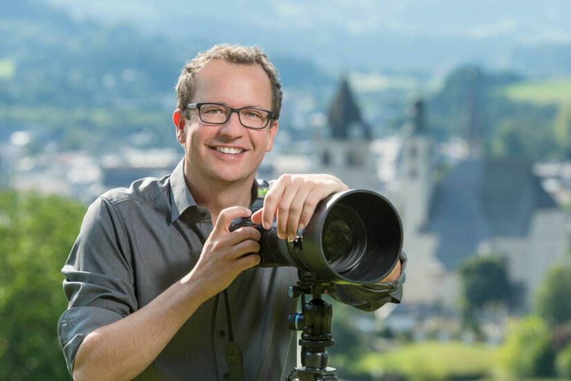 Ein lächelnder Fotograf mit einer Kamera steht im Freien. Im Hintergrund sind unscharfe Gebäude und eine grüne Landschaft zu sehen.