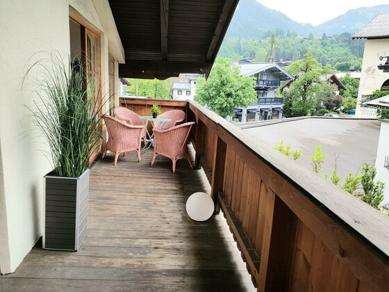 Holzbalkon mit rosa Korbsesseln, einem kleinen Tisch und Blick auf alpine Gebäude und bewaldete Berge.